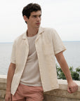 Man wearing a beige shirt and pink shorts standing by a body of water.