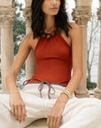 Woman wearing a rust-colored halter top and white pants sitting outdoors.