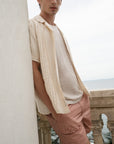 Person wearing a beige shirt and pink pants leaning against a wall with a scenic background.