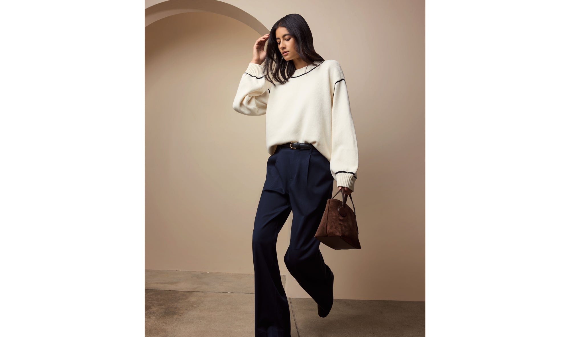 Woman in a white sweater and dark pants holding a brown bag against a beige wall.