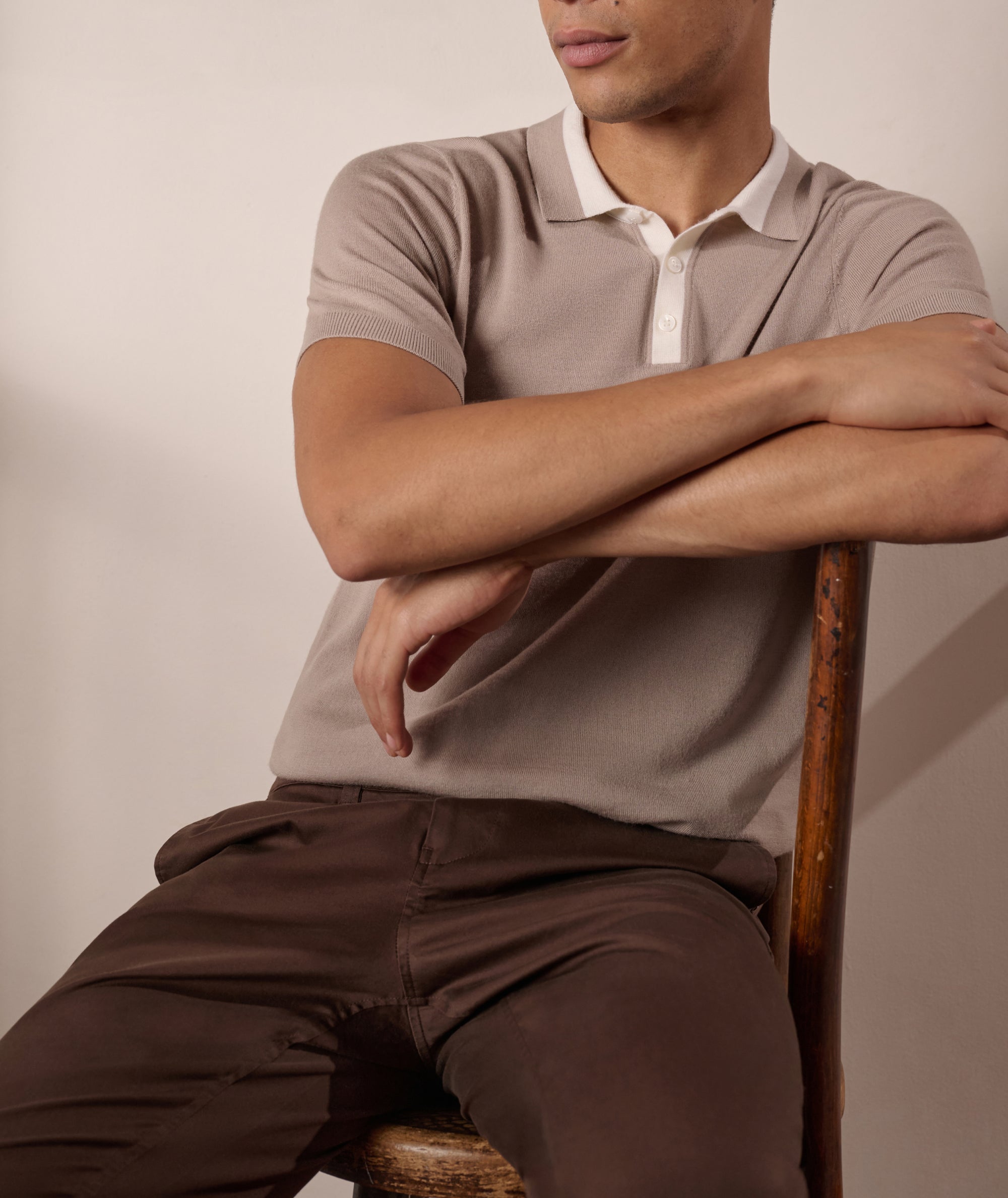 Man sitting on a chair wearing a beige polo shirt and brown pants against a plain background
