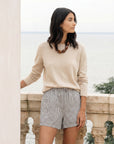 Woman wearing a beige sweater and striped shorts standing on a stone balcony with a scenic background.