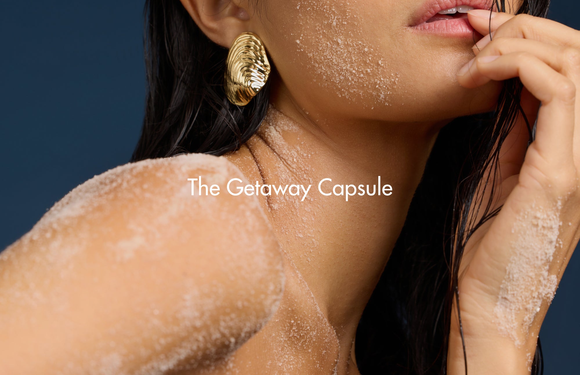 Woman with gold earrings and sand on her skin, with 'The Getaway Capsule' text.