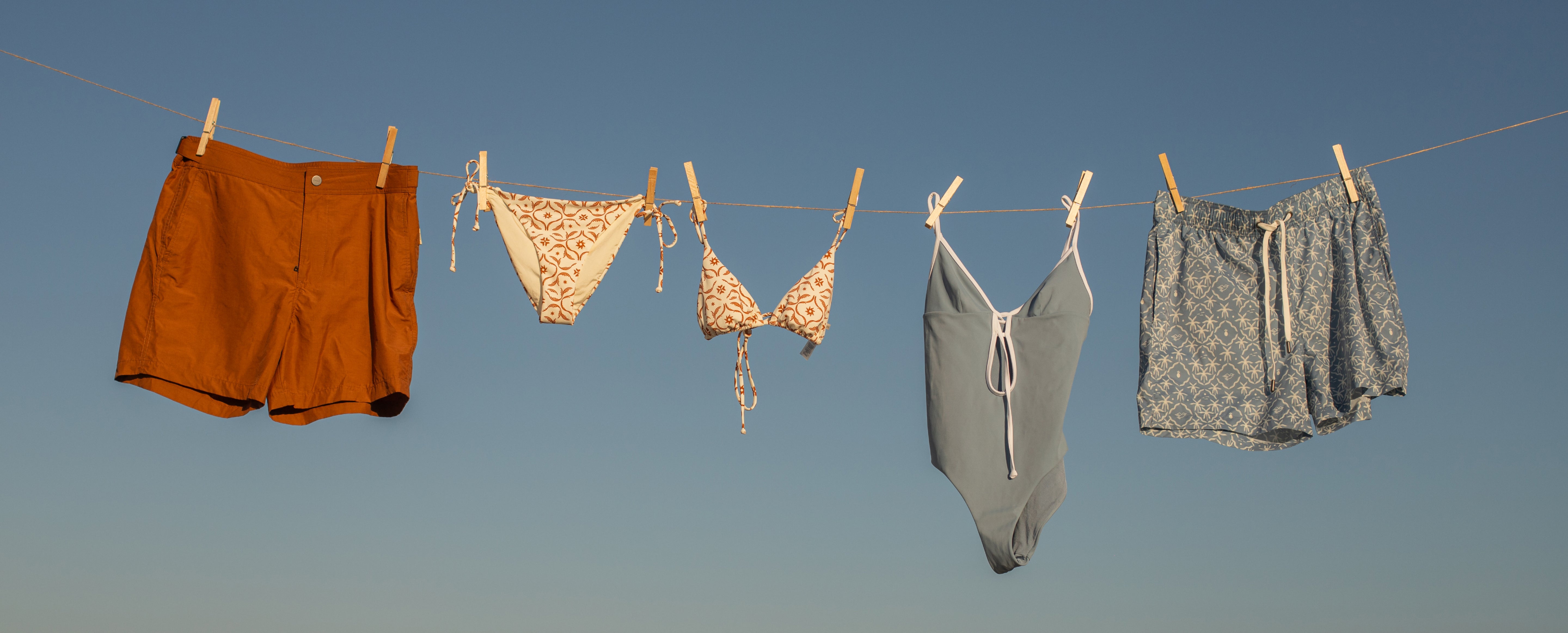 Clothing items including shorts, a bikini, and a swimsuit hanging on a clothesline against a clear blue sky.
