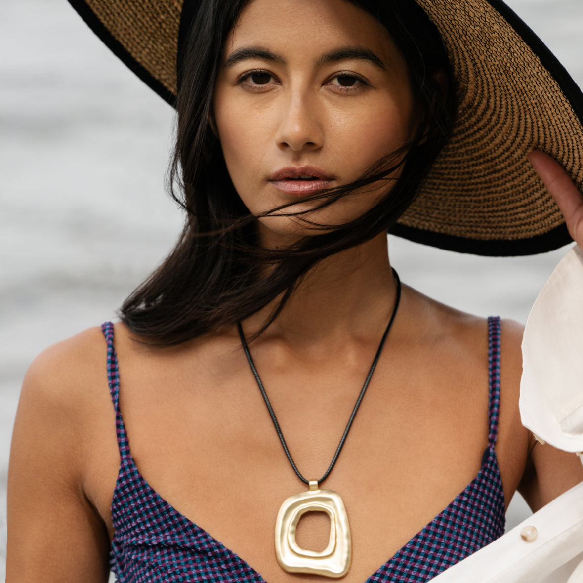 Woman wearing a large gold pendant necklace and a wide-brimmed hat against a neutral background